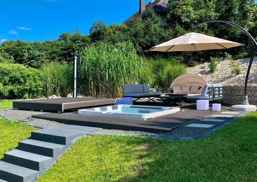 Whirlpool, nahezu bündig in eine dunkle Holzterrasse eingelassen, um die Terrasse Kies, dahinter Gräser, Lounge-Möbel und ein Sonnenschirm