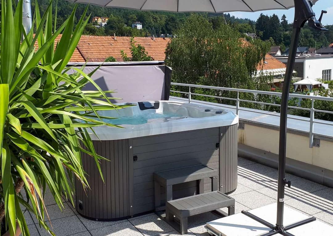 Freistehender Whirlpool mit grauer Verkleidung und Einstiegstreppe auf einer Dachterrasse, daneben ein Sonnenschirm