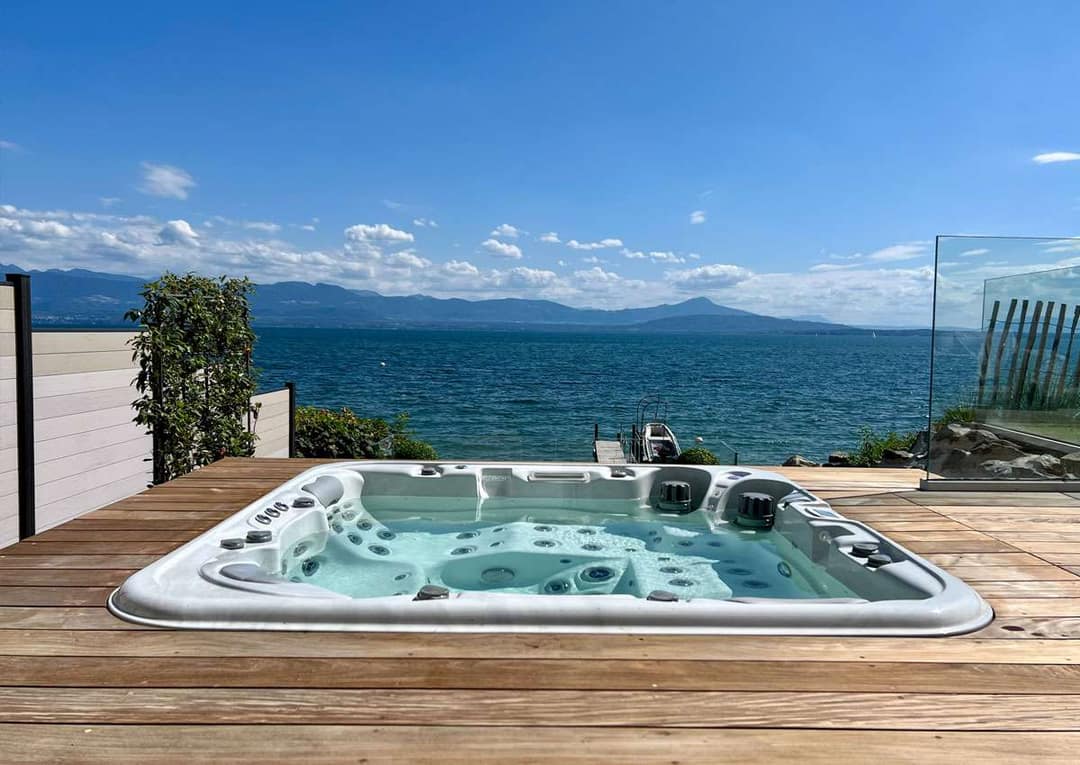 In eine Holzterrasse eingelassener Whirlpool, dahinter ein See und eine Bergkette