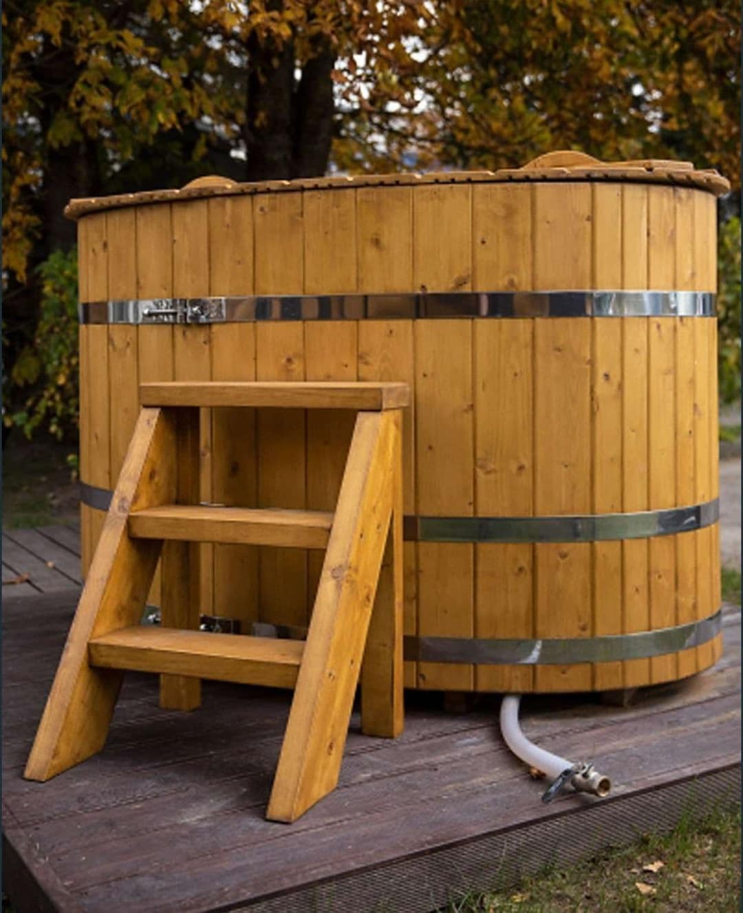 Ovale Holzbadewanne auf einem Holzdeck, davor eine Holztreppe, unten ein Schlauch am Ablassventil