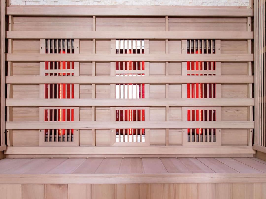 Rückwand einer Infrarotkabine aus hellem Holz mit rot glühenden Strahlern hinter Holzgittern, davor eine Bank