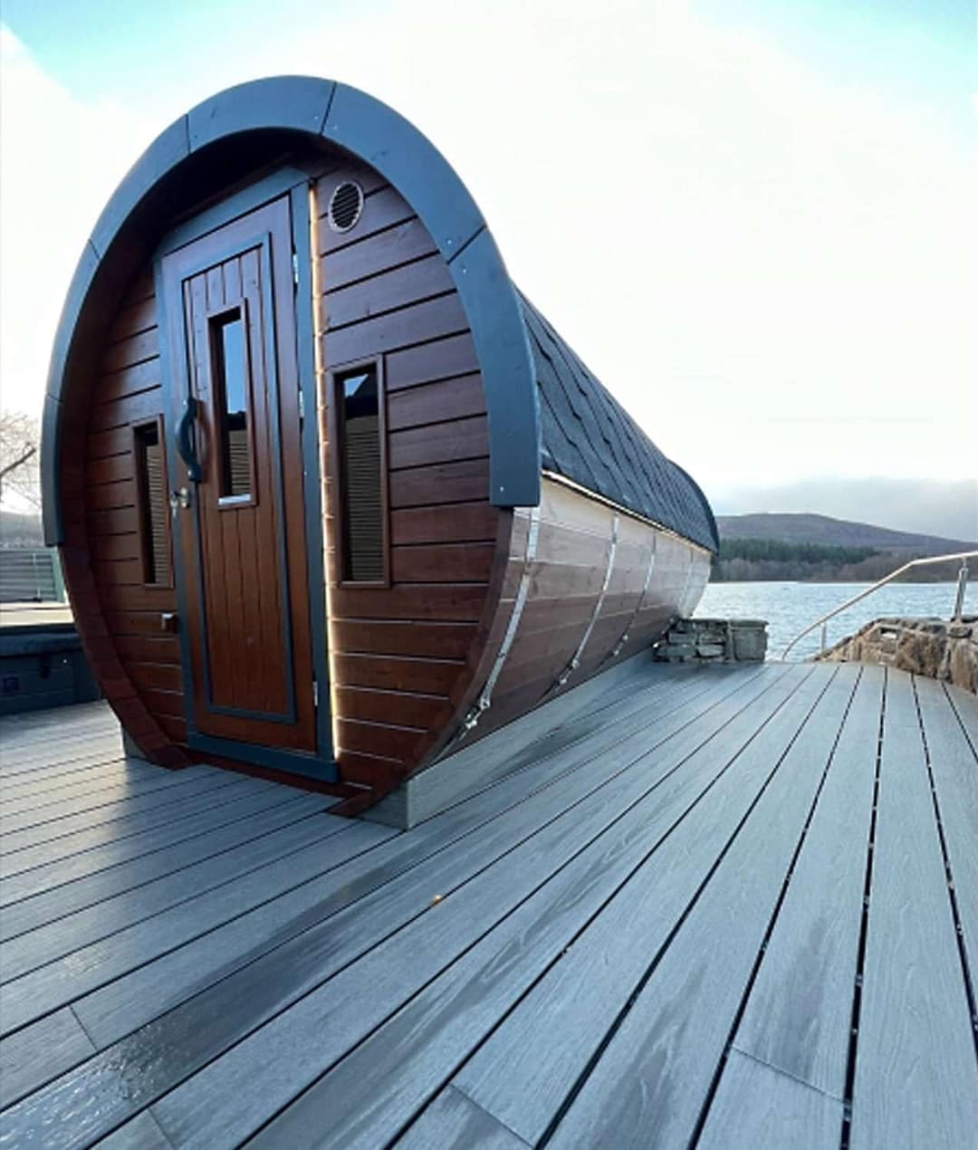 Saunafass aus dunklem Holz auf einem Holzdeck an einem See