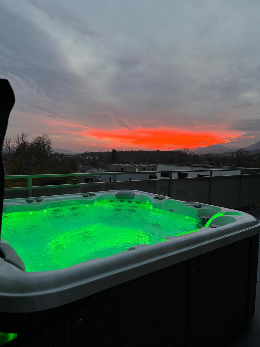 Whirlpool mit grün beleuchtetem Wasser auf einer Dachterrasse mit Geländer, am Abendhimmel ein roter Streifen