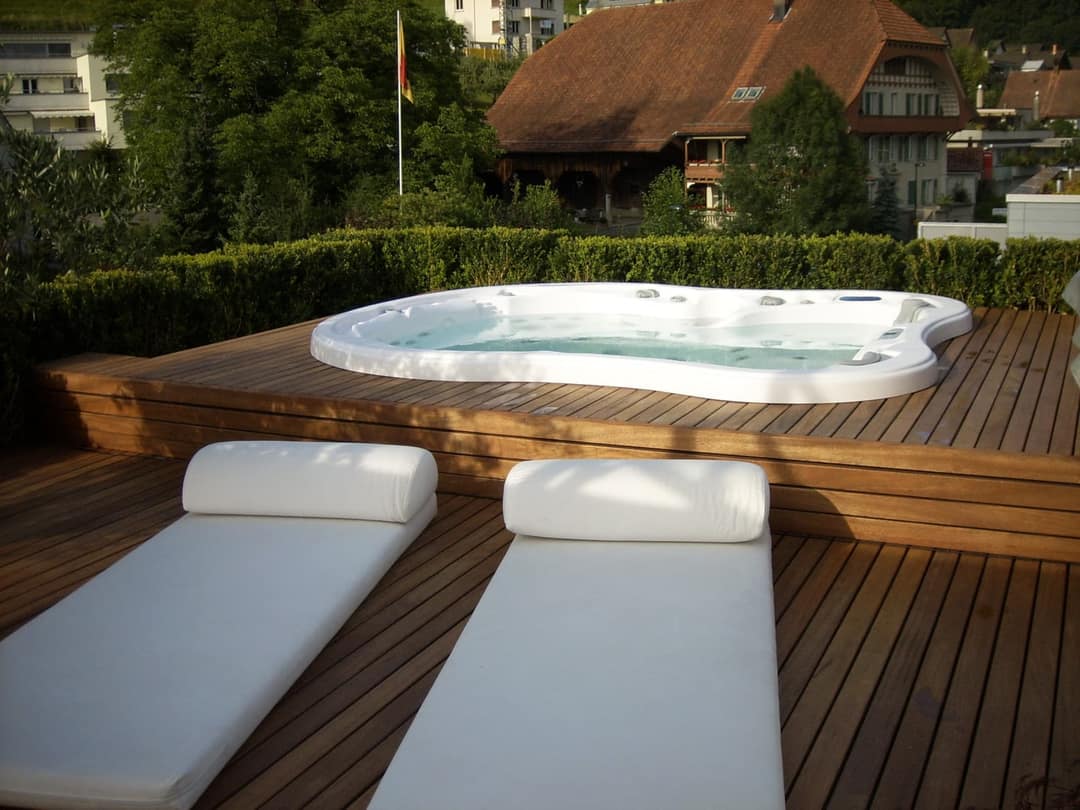 Whirlpool, bündig in eine erhöhte Holzterrasse eingelassen, davor zwei weisse Liegen, dahinter eine Hecke und ein Bauernhaus