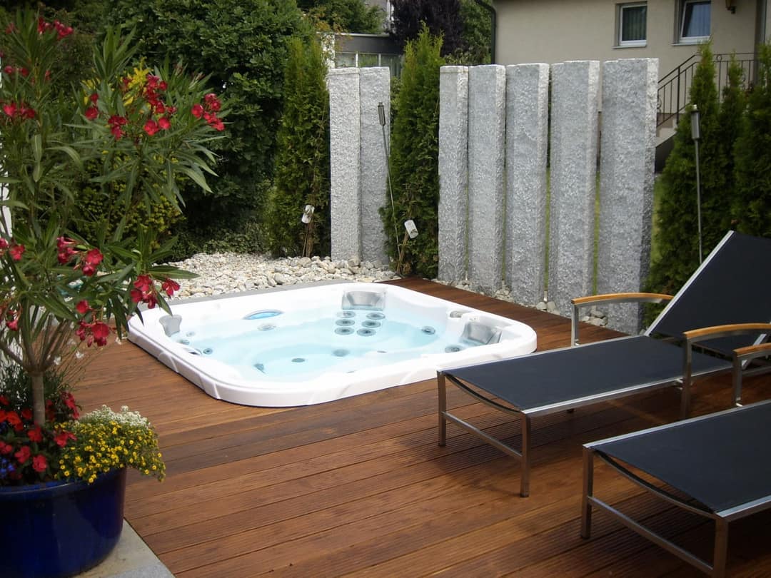 Whirlpool, bodeneben in eine Holzterrasse eingelassen, dahinter Granitstelen und Thujahecken, rechts zwei Liegen