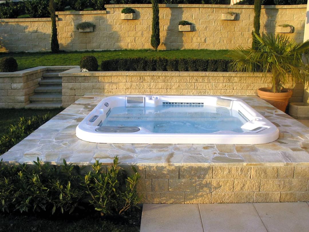 Whirlpool, bündig in eine Einfassung aus Natursteinplatten eingelassen, dahinter ein Garten mit Natursteinmauern und einer Treppe