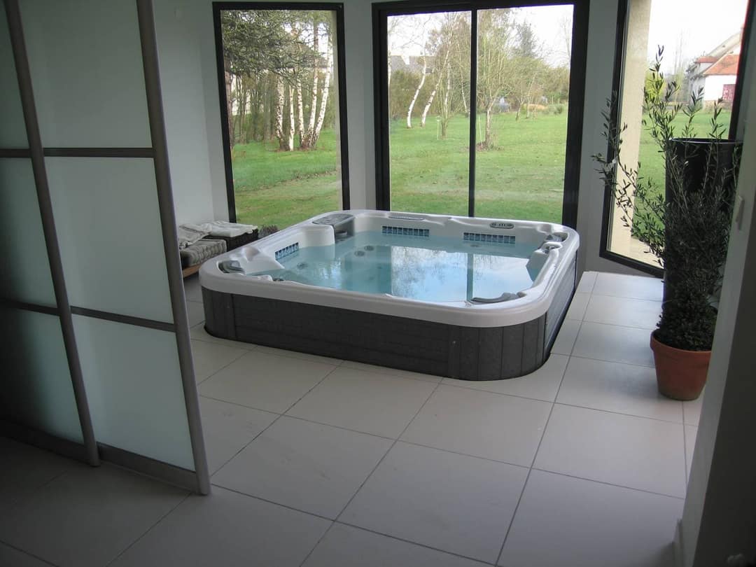 Teilversenkter Whirlpool mit grauer Verkleidung in einem Raum mit Fliesenboden und bodentiefen Fenstern zum Garten